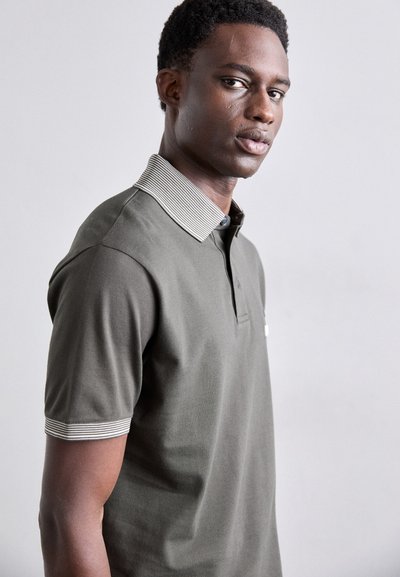 Olivza zaļa polo krekls ar svītrotu apkakli, īsām piedurknēm un smalku tekstūru. Iekļauj pogu priekšu un mazu logotipu.