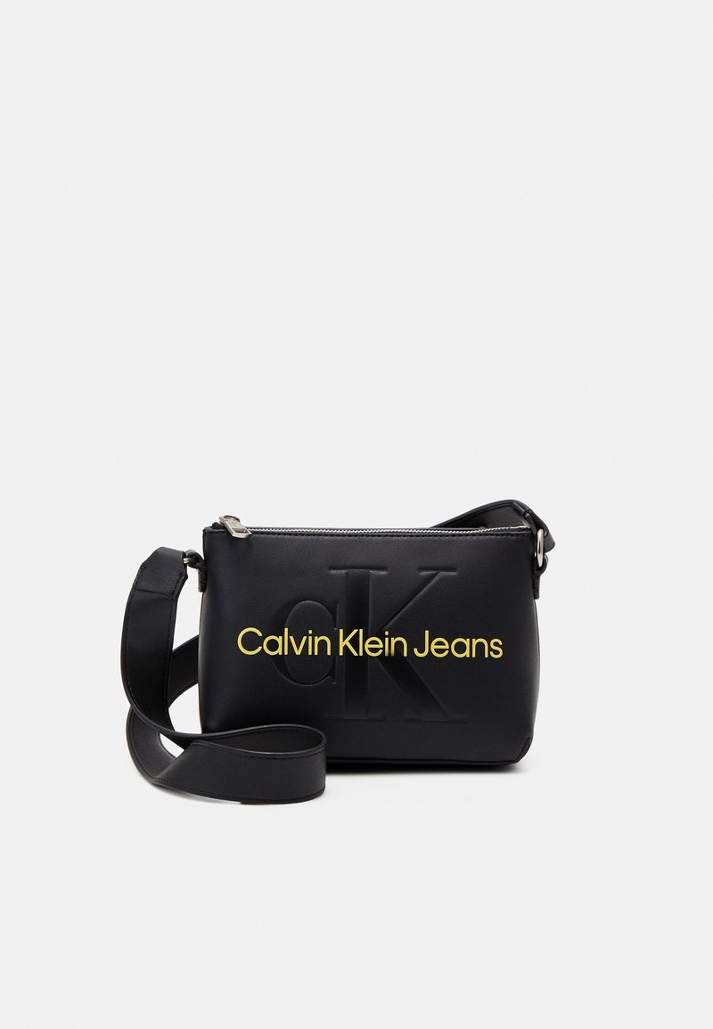 Musta olkalaukku, joka on valmistettu sileästä nahasta, jossa on kohokuvioitu 'cK' ja keltainen 'Calvin Klein Jeans' -logo. Laukussa on säädettävä hihna ja vetoketjClosure.