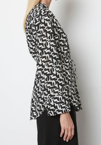 Blusa a maniche lunghe in bianco e nero con design a incrocio, legata in vita, caratterizzata da una stampa geometrica e una texture di tessuto liscia.
