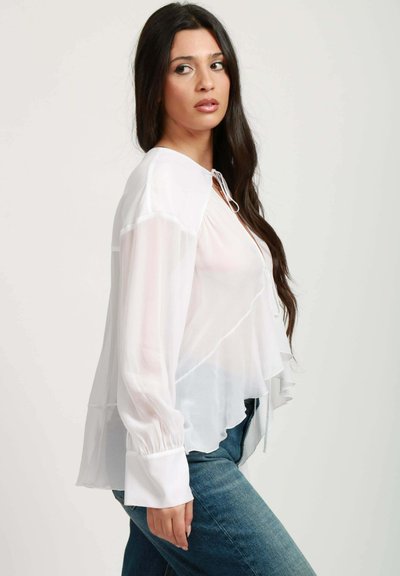Blusa bianca trasparente con orlo arricciato, collo tondo e dettaglio con cordoncino. Maniche lunghe con polsini arricciati, design a strati e tessuto leggero.