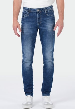 Mann trägt slim-fit blaue verwaschene Jeans mit Vordertaschen und weißen Sneakers, steht vor einem schlichten hellen Hintergrund.