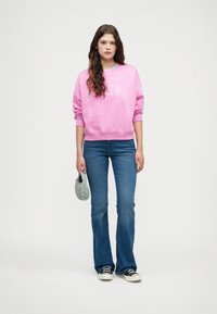 Sweatshirt rose clair avec texte blanc, associé à un jean évasé bleu et des baskets noires. La tenue présente une coupe décontractée et une texture intermédiaire.
