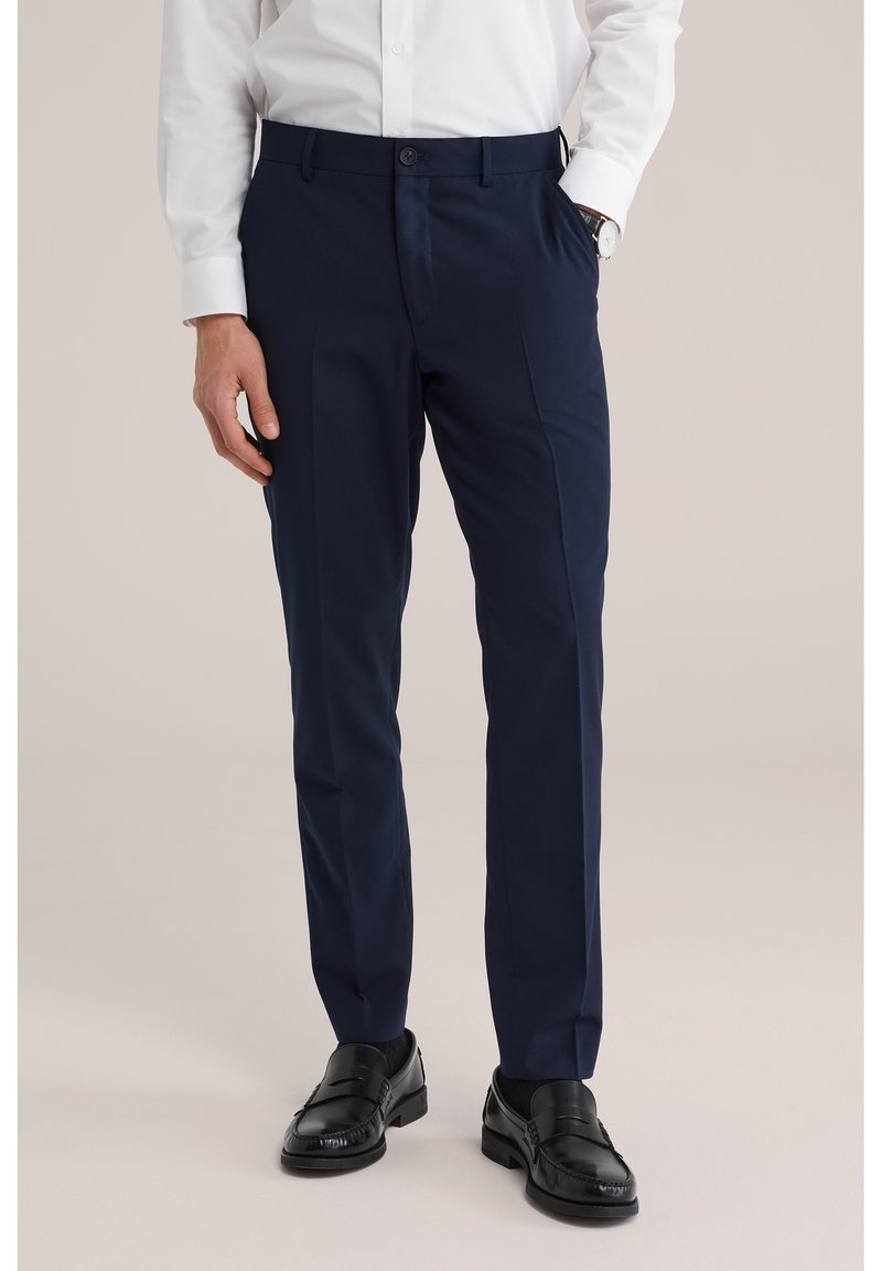 Mann trägt marineblaue Anzughose, schwarze Anzugschuhe und ein weißes langärmliges Hemd mit Knöpfen, eine Hand in der Tasche.