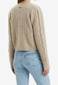 Beige svetr s pleteným vzorem a žebrovým lemem. V kombinaci se světle modrými džíny s zadními kapsami a červenou nášivkou Levi's.