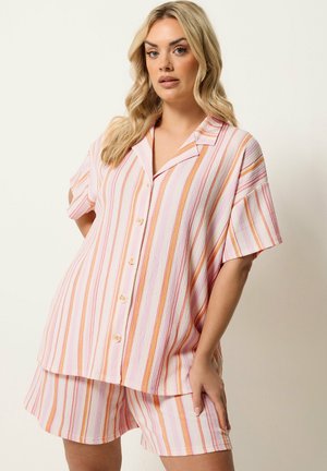 Kobieta pozująca w koszuli z krótkim rękawem na guziki oraz dopasowanych szortach z pionowymi paskami w kolorach różowym, pomarańczowym i białym na jasnym tle.