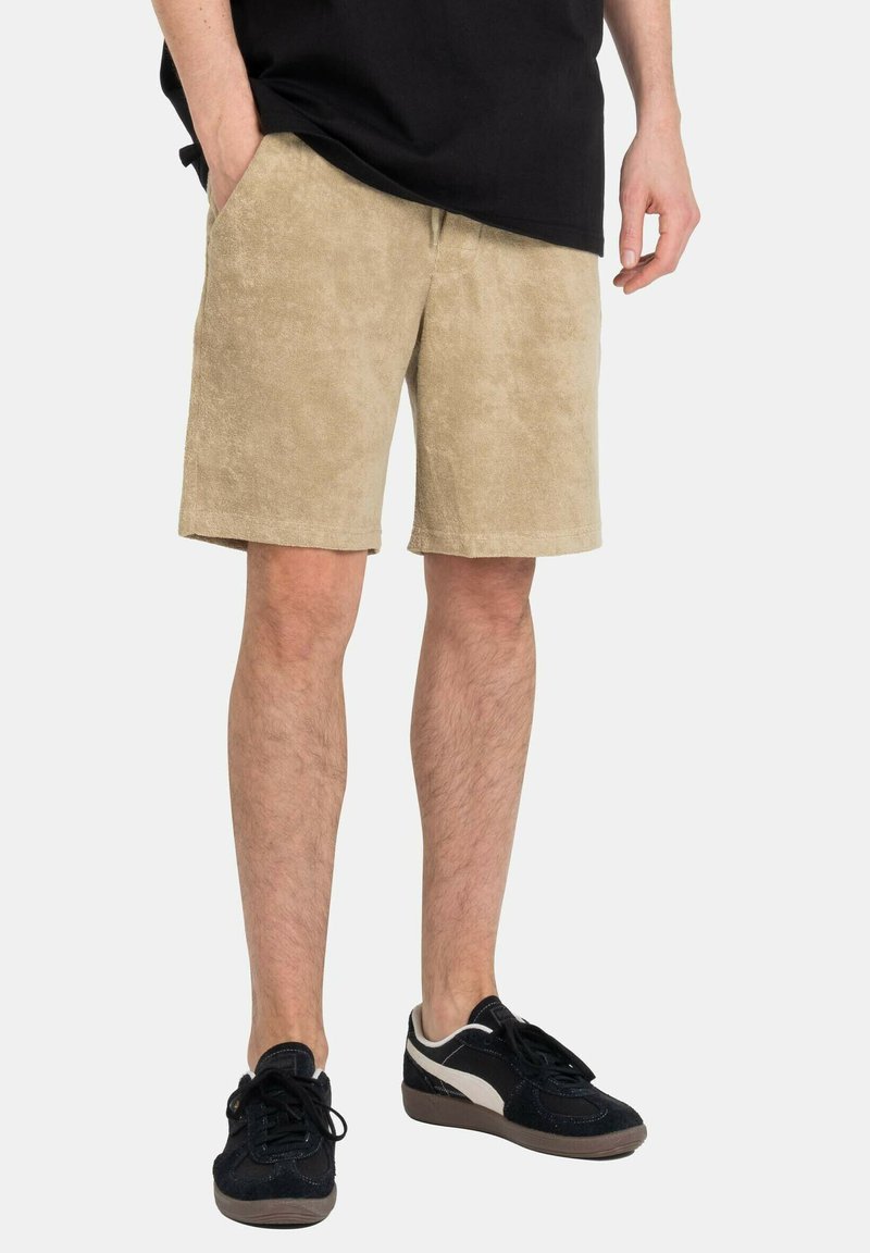 Beige, strukturierte Shorts aus einem weichen Stoff. Sie verfügen über zwei Seitentaschen und einen elastischen Bund, kombiniert mit schwarzen Turnschuhen.
