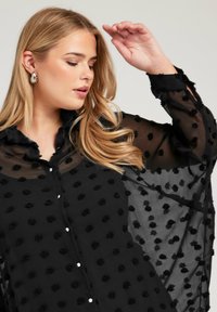 Svart genomskinlig blus med texturerad blommig applikation, knäppning fram och flödande ärmar. Har en klassisk krage och mjukt, delikat tyg.