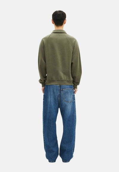 Man staat met zijn rug naar de kijker, gekleed in een olijfgroen sweatshirt en losse blauwe spijkerbroek tegen een effen witte achtergrond.