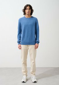 Blå ulltröja med rundad halsringning, ribbade manschetter och nederkant, kombinerad med beige smalbyxor och vita sneakers, mot en neutral bakgrund.