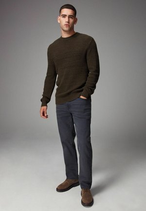 Olive textured knit sweater, gray straight-leg pants, and brown suede lace-up shoes. Model standing with one hand in pocket, neutral background.