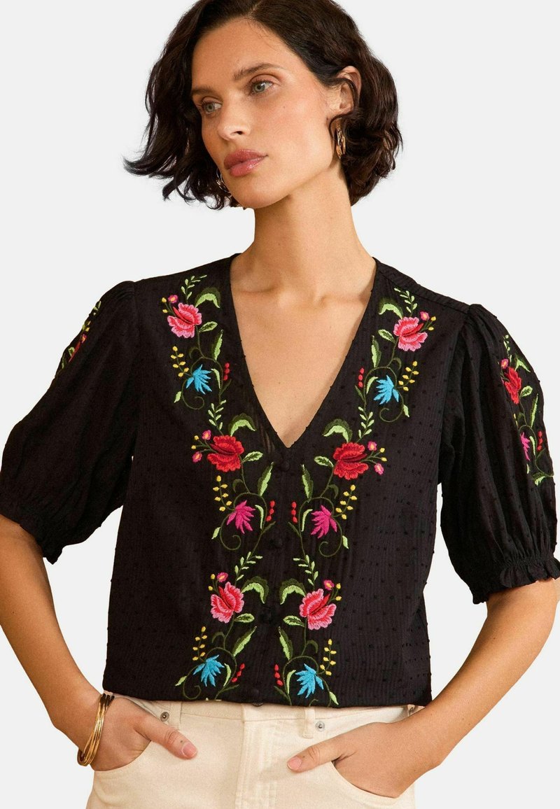 Fekete blúz puffos ujjakkal, mély V-kivágással és színes rózsaszín és kék virágmintázattal. Textúrált anyag gombos díszítéssel.
