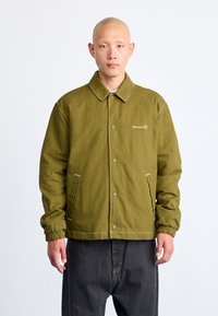 Veste vert olive avec un col, des boutons à pression, des poignets élastiques et un logo Timberland. Fabriquée dans un matériau robuste avec des coutures contrastées.
