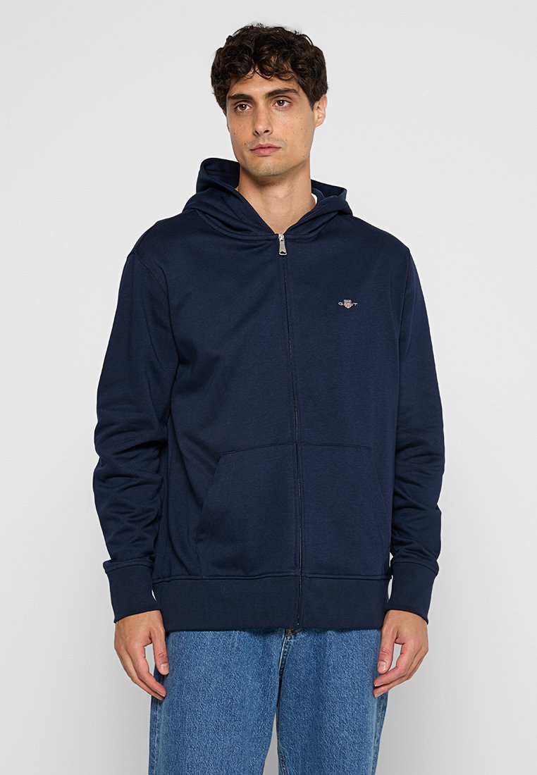 Marinblå zip-up hoodie gjord av mjukt tyg, med en framficka, justerbar huva och en liten broderad logotyp på bröstet.