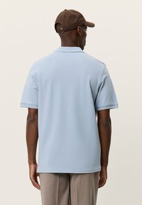 Ανοιχτό μπλε πόλο με κοντά μανίκια, κατασκευασμένο από απαλό ύφασμα, με κλασικό γιακά. Φορεμένο με καφέ καπέλο και μπεζ παντελόνι.