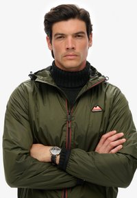 Olivgrön vattentät jacka med blixtlås framtill, svart fleece-krage och röda detaljer. Har en logotyp patch på bröstet samt justerbar huva.