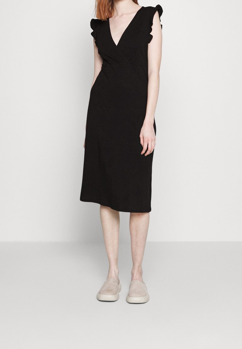 Robe noire avec un col en V, manches courtes à volants et une silhouette ajustée. Tissu lisse, longueur genou, associée à des chaussures beige sans lacets.