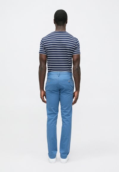 Ριγέ μπλε μπλουζάκι και ανοιχτόχρωμο μπλε παντελόνι, που τονίζει μια λεπτή εφαρμογή. Το παντελόνι έχει πίσω τσέπες με κουμπιά. Φαίνονται λευκά αθλητικά παπούτσια.