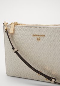 Borsa a tracolla bianca texture con hardware dorato, dotata di un logo, chiusura con zip e una tracolla regolabile marrone.