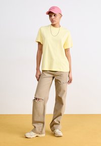 T-shirt en coton jaune, coupe décontractée, associé à un jean beige baggy avec des accros. Une casquette rose et des baskets beiges complètent la tenue.
