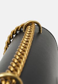 Sac à main en cuir noir avec une épaisse bandoulière en chaîne dorée et du matériel circulaire doré embossé de "PINKO". Texture lisse avec un design minimaliste.
