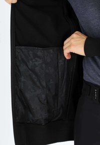 Black zippered jacket with a textured interior pocket featuring a dark camouflage pattern. The cuffs and hem are ribbed for fit.