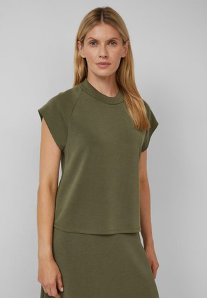 Frau mit langen blonden Haaren, die ein olivgrünes ärmelloses Oberteil und einen passenden Rock trägt, steht vor einem schlichten grauen Hintergrund.