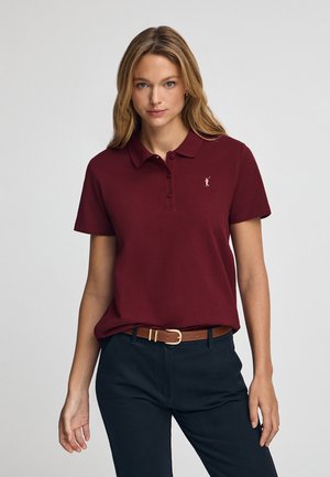 Polo shirt bourgogne avec col et trois boutons, arborant un petit logo doré sur la poitrine gauche. Porté avec un pantalon marine et une ceinture marron.
