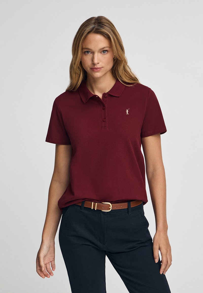 Bordeaux poloshirt met een kraag en drie knopen, voorzien van een klein gouden logo op de linkerborst. Draag het met een marineblauwe broek en een leren bruine riem.