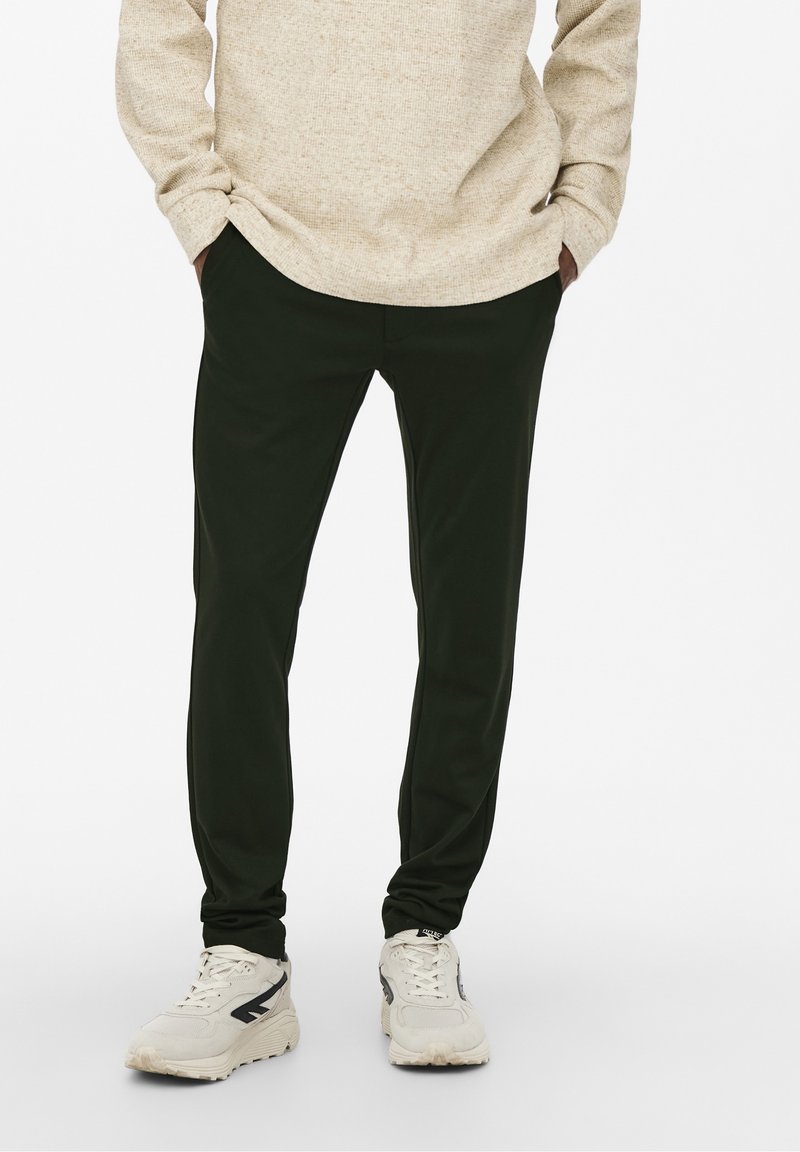 Dark green, slim-fit pants with a smooth texture and side pockets, paired with light gray athletic sneakers featuring black accents.