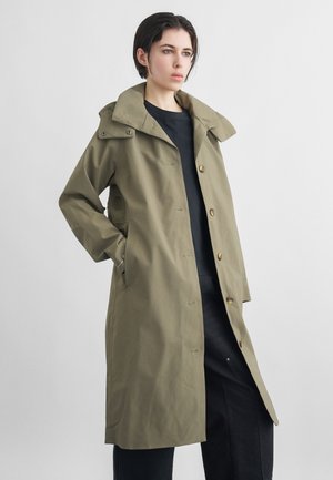 Frau mit kurzem dunklem Haar, die einen langen olivgrünen Mantel mit Knöpfen über einem schwarzen Hemd und einer schwarzen Hose trägt, stehend mit einer Hand in der Tasche.