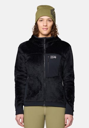 Personne portant une veste polaire noire avec une poche poitrine et un bonnet vert olive, debout devant un fond gris uni.