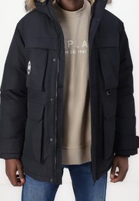 Svart vadderad parka med en huva fodrad med päls, flera framfickor och dragkedja, lager över en beige sweatshirt och denimjeans.