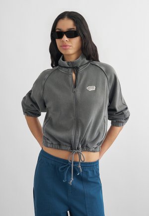 Młoda kobieta w czarnych okularach przeciwsłonecznych, ubrana w szarą kurtkę zapinaną na zamek z elastycznym ściągaczem i niebieskie spodnie z sznurkiem, stojąca z rękami za plecami.