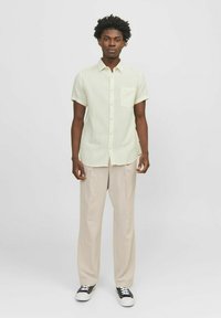 Chemise jaune clair à manches courtes avec une poche poitrine, associée à un pantalon beige et des baskets noires. Tissu lisse et coupe décontractée.
