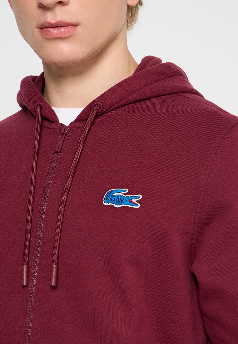 Haut à zip de couleur bordeaux en mélange de coton, avec un logo de crocodile brodé bleu sur la poitrine et des cordons de serrage sur la capuche.