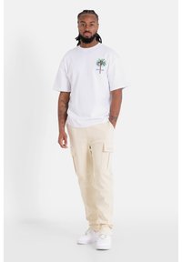 Witte t-shirt met korte mouwen en een palmboomafbeelding op de linkerkant van de borst, gecombineerd met beige cargobroek met zijzakken en witte sneakers.