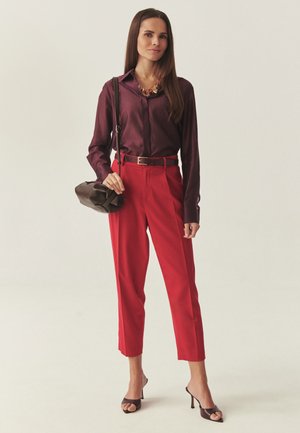 Mujer de pie con una blusa burdeos, pantalones rojos de talle alto, sandalias marrones con tacón, lleva un bolso marrón al hombro y un collar dorado llamativo.