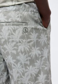 Gros plan sur un short gris à imprimé de palmiers blancs, poche arrière et un logo circulaire "B" près de la poche, main reposant à l'intérieur de la poche latérale.