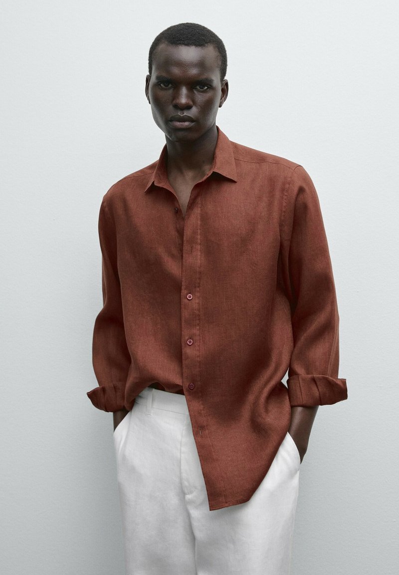 Rust-colored linen shirt with long sleeves, buttoned front, and rolled cuffs, paired with white trousers. Smooth texture, minimal design.