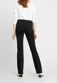 Mujer con una camisa blanca de manga larga y jeans negros, vista desde atrás. Ella está de pie contra un fondo claro y liso.