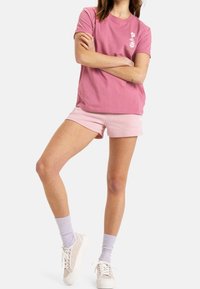 Žena v růžovém tričku s květinovým potiskem a svetle růžových šortkách, stojící s překříženýma rukama a s jednou nohou mírně zdviženou.