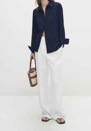 Chemise bleu marine à boutons avec manches longues, pantalon large à rayures blanches, sac panier tressé avec des accents marron, assortis à des tongs noires.