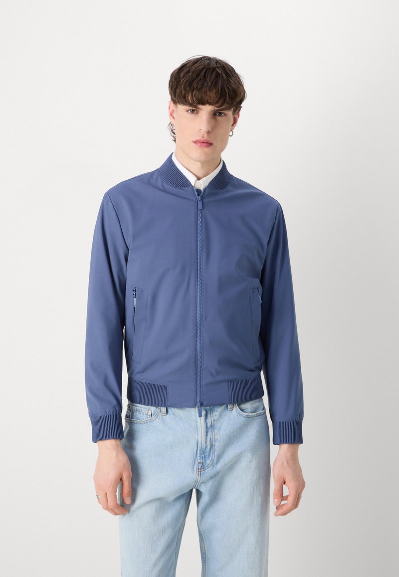 Veste bomber bleue avec zip à l'avant, col et poignets côtelés, dotée de deux poches latérales, fabriquée en tissu lisse. Portée sur un jean clair.