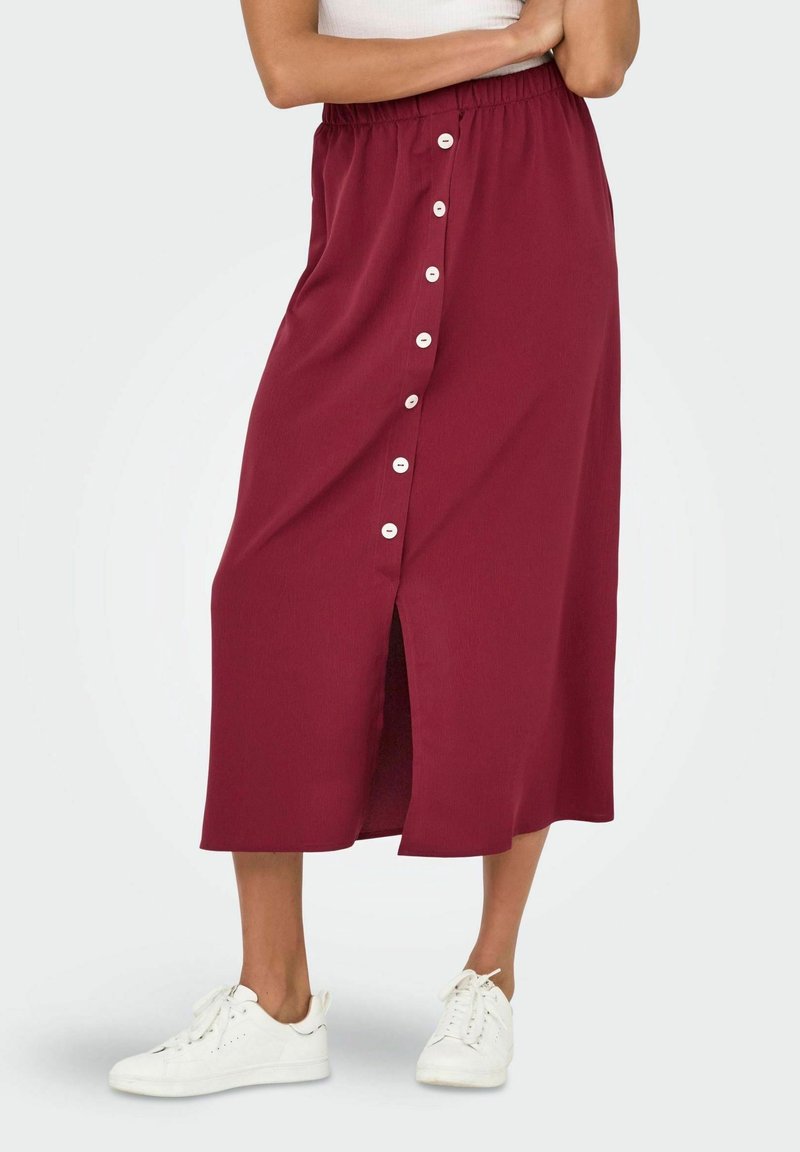 Femme portant une jupe midi bordeaux avec des boutons sur le devant et des baskets blanches, debout les bras croisés sur un top blanc.