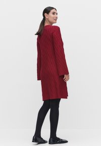 Röd stickad kappa med diamantmönster, långa ärmar och en något utställd fåll, kombinerad med svarta strumpbyxor och ballerina-skor. Neutral bakgrund.