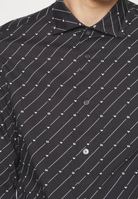 Czarna koszula z diagonalnym wzorem białych linii i małymi logotypami EA. Ma kołnierzyk zapinany na guziki oraz standardowe guziki na przodzie.