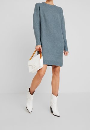 Pletené svetrové šaty v modré barvě s žebrovanou texturou a dlouhými rukávy. Kombinované s bílou kabelkou a bílými kovbojskými botami s špičatou špičkou.