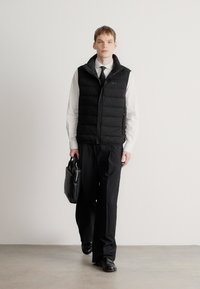 Μαύρο γιλέκο puffer με φερμουάρ, πάνω από λευκό πουκάμισο και μαύρο παντελόνι. Μαύρη δερμάτινη τσάντα στο χέρι. Το μοντέλο με κοντά μαλλιά στέκεται μπροστά σε λευκό φόντο.