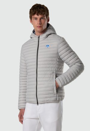 Uomo che indossa una giacca imbottita grigio chiaro con cappuccio, cerniera nera e logo circolare blu, abbinata a pantaloni bianchi, in piedi contro uno sfondo semplice.