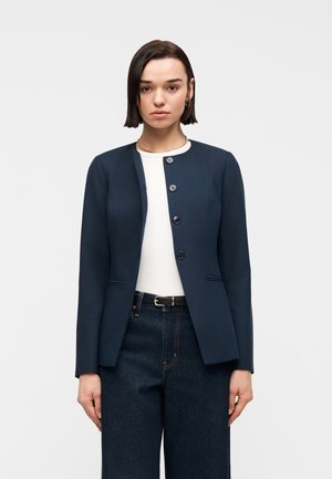 Vrouw draagt een donkerblauwe gebuttonde blazer over een wit topje en donkere jeans met hoge taille en een zwarte riem, staand tegen een effen lichte achtergrond.